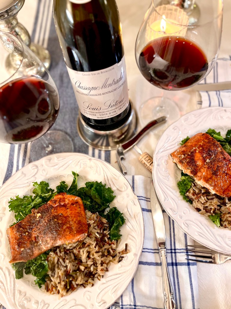 Two plates of blackened salmon laid over a bed of citrus wild rice and kale. Also on the table, two glasses of red wine and a bottle of Louis Latour 2005 Pinot Noir.